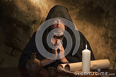 portrait of an elderly monk in a black robe, hands folded in prayer, a dark cell, a candle burning on a table. Stock Photo