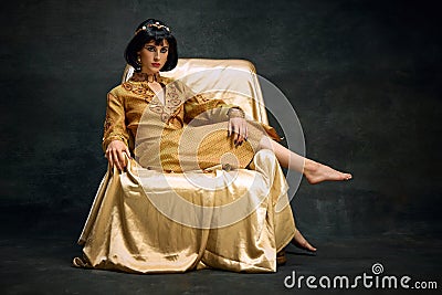 Portrait of elegant, beautiful woman in image of antique queen, Cleopatra in olden clothes sitting on armchair against Stock Photo