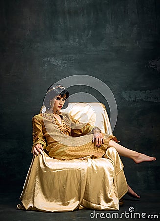 Portrait of elegant, beautiful woman in image of antique queen, Cleopatra in olden clothes sitting on armchair against Stock Photo