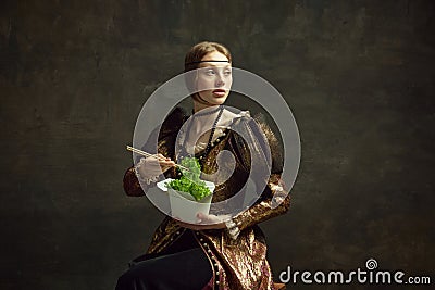 Portrait of elegant young girl, queen in vintage dress eating lettuce with chopstick against dark green background Stock Photo