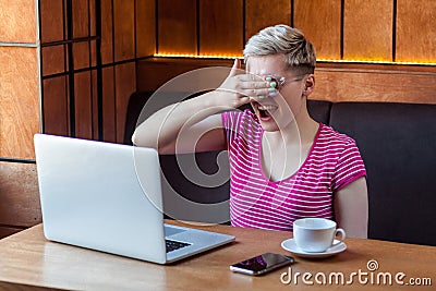 Portrait of emotional scared happy young businesswoman in pink t-shirt is sitting in cafe and covering the eyes to herself with Stock Photo