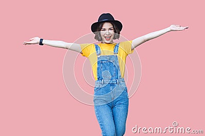 Portrait of excited beautiful young woman in yellow t-shirt, blue denim overalls, makeup, black hat standing with raised arms and Stock Photo