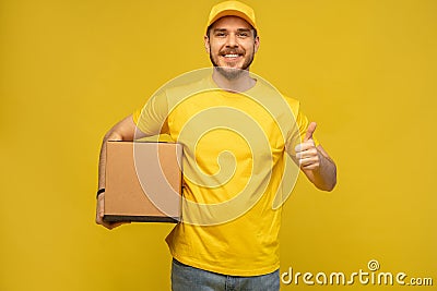 Portrait of excited delivery man in yellow uniform holding paper box isolated over yellow background. Stock Photo