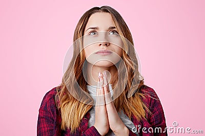 Portrait of faithful adorable female keeps hands together, asks God for wellbeing and health, looks hopefully upwards, isolated ov Stock Photo