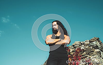 Portrait of fashion man model wearing dark clothes posing on blue sky background. Young strong muscular man standing on Stock Photo