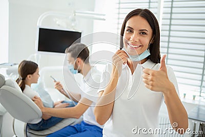 Portrait of female dentist showing thumb up while her collegue working with small client on the background Stock Photo