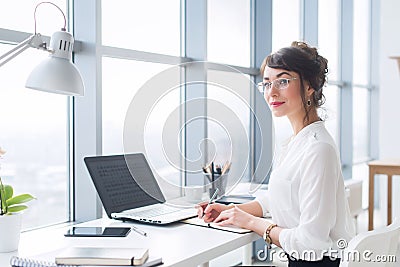 Portrait of a female writer working at office, using laptop, wearing glasses. Young employee planning her work day Stock Photo