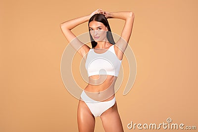 Portrait of fit woman in white lingerie posing on beige studio background, raising arms up, showing smooth armpits Stock Photo