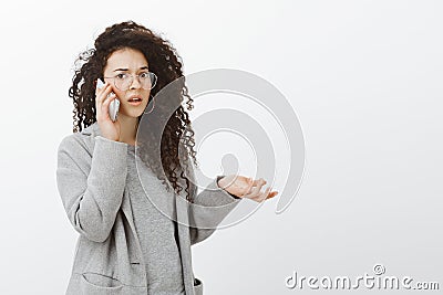 Portrait of frustrated questioned girlfriend with curly hair in grey coat and glasses, raising hand cluelessly while Stock Photo