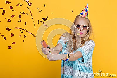 Portrait of funny blond woman in birthday hat and red confetti on yellow background. Celebration and party. Stock Photo