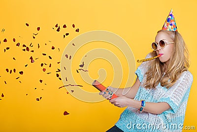 Portrait of funny blond woman in birthday hat and red confetti on yellow background. Celebration and party. Stock Photo