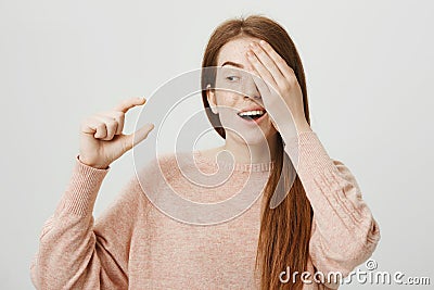 Portrait of funny emotive ginger girl covering one eye while showing something small or tiny with gesture and being Stock Photo