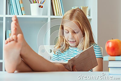 Portrait of funny little school girl relaxing reads book at the table in room at home Stock Photo