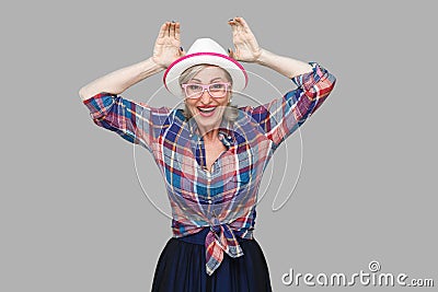 Portrait of funny modern stylish mature woman in casual style with hat and eyeglasses standing with bunny ears gesture and looking Stock Photo
