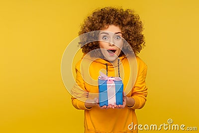 Portrait of funny surprised curly-haired young woman in urban style hoodie holding blue gift box Stock Photo