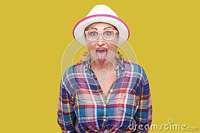 Portrait of funny surprised modern stylish mature woman in casual style with hat and eyeglasses standing tongue out, looking at Stock Photo