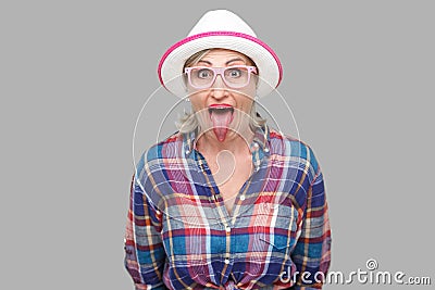 Portrait of funny surprised modern stylish mature woman in casual style with hat and eyeglasses standing tongue out, looking at Stock Photo