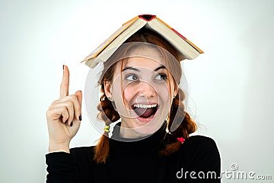 Portrait of funny young smiling student girl with an open book on her head holding her point finger up having an idea. Reading and Stock Photo