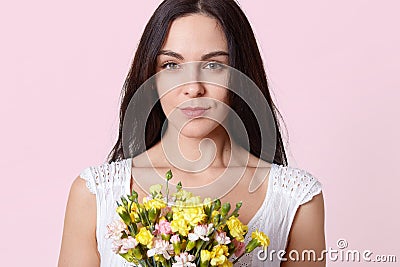 Portrait of gentle brunette young lady, dressed in white dress, recieves gift on birthday, being photographed with serious facial Stock Photo