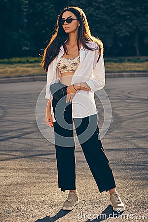 Portrait of a girl in dark sunglasses posing in city. Dressed in top with floral print, white shirt, black trousers Stock Photo