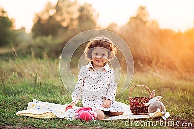 Portrait of a girl on a meadow in the sunset sunlight. Little girl in a light overalls sits on a blanket next to basket with harve Stock Photo