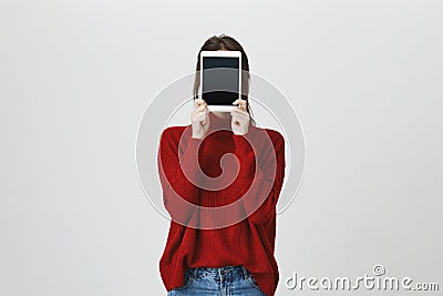 Portrait of girl in trendy red sweater covering her face with a tablet standing near white wall. Technology and gadget Stock Photo