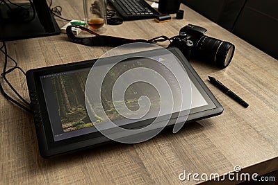 A portrait of a graphical drawing pen tablet and its stylus lying on a wooden table next to a photo camera. The tablet is Stock Photo