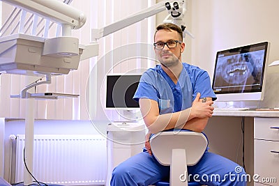 Portrait of a handsome bearded dentist male in glasses wearing a blue uniform, sitting in a dentist office. Stock Photo