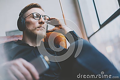 Portrait handsome bearded man glasses,headphones listening to music modern loft studio.Man sitting in vintage chair,holding smartp Stock Photo