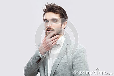 Portrait of handsome brown bearded serious man in white shirt and casual grey suit standing, touching his face and looking at Stock Photo