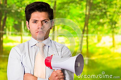 Portrait of a handsome mad man holding in his hand a megaphone with his arms crossed, in a blurred green background Stock Photo