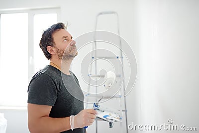 Portrait of a handsome mature man making repairs in the apartment. The person paints the wall white with roller. Do it yourself. Stock Photo