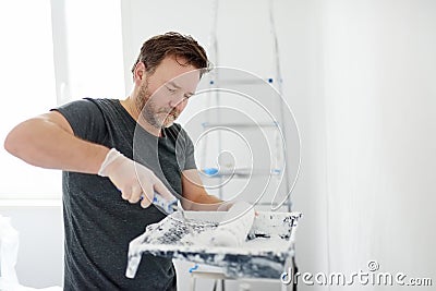 Portrait of a handsome mature man making repairs in the apartment. The person paints the wall white with roller. Do it yourself. Stock Photo