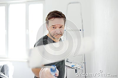 Portrait of a handsome mature man making repairs in the apartment. The person paints the wall white with roller. Do it yourself. Stock Photo