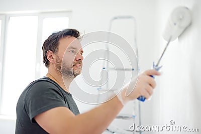 Portrait of a handsome mature man making repairs in the apartment. The person paints the wall white with roller. Do it yourself. Stock Photo