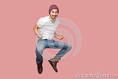 Portrait of happy bearded hipster young man in white shirt and blue jeans with hat jumping, rejoicing and celebrating his victory Stock Photo