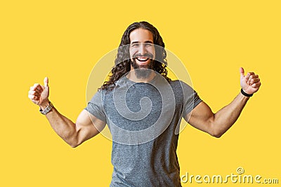 Portrait of happy bearded young man with long curly hair in grey tshirt standing with thumbs up and looking at camera with toothy Stock Photo