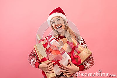 Portrait of happy blonde woman in cozy sweater and Santa hat holding heap of Christmas presents on pink background Stock Photo