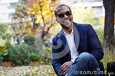 Portrait of Happy confident young African-American businessman in formal wear and sunglasses sitting at city park Stock Photo