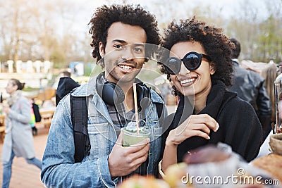 Portrait of happy cute dark-skinned couple with afro hairstyle, strolling on food festival, tasting and drinking Stock Photo