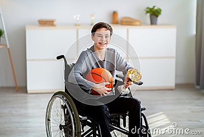 Portrait of happy disabled teen in wheelchair holding basketball and prize, smiling at camera indoors Stock Photo