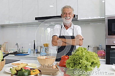 Portrait of happy elderly man with apron standing with arms crosse at kitchen with colorful fresh vegetables, fruits,ingredients. Stock Photo