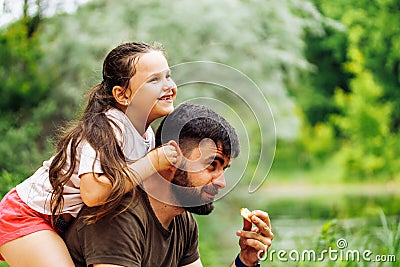 Portrait of happy family sitting on picnic in park forest around trees bushes. Little daughter sitting on fathers back. Stock Photo