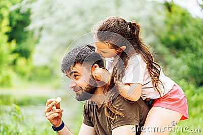 Portrait of happy family sitting on picnic in park forest around trees bushes. Little daughter sitting on fathers back. Stock Photo