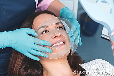 Portrait of happy female patient sitting in dentist chair, smiling and examining her teeth in the mirror, dental surgeon showing Stock Photo