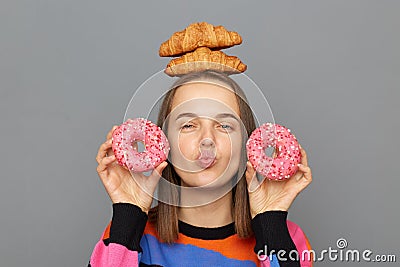 Portrait of happy joyful woman in sweater holding donuts in hands, standing with croissant on her head, looking at camera with Stock Photo