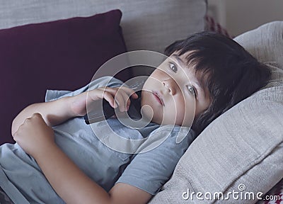 Portrait of happy kid lying on sofa watching TV, Positive child boy holding remote control looking up with smilng face. Relaxing, Stock Photo