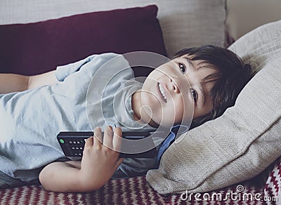 Portrait of happy kid lying on sofa watching TV, Positive child boy holding remote control looking up with smilng face. Relaxing, Stock Photo