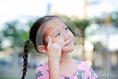 Portrait of happy little Asian child in garden with thinking and looking up. Close-up smiling kid girl with forefinger point on Stock Photo