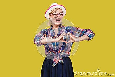 Portrait of happy peaceful modern stylish mature woman in casual style with white hat standing with hand heart shape gesture, Stock Photo